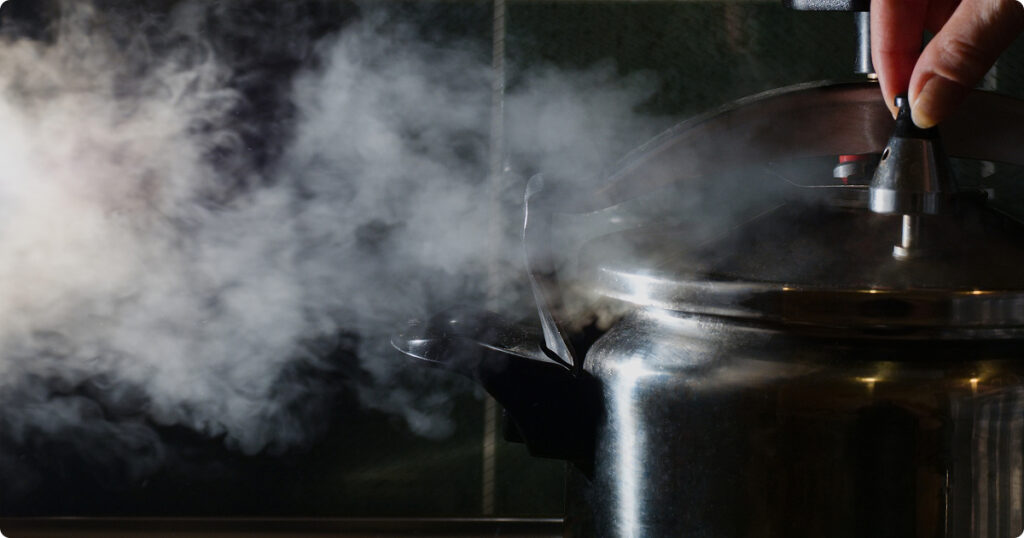 Steam erupting from a stainless steel pressure cooker as a person releases the valve, symbolizing the danger of defective pressure cookers.