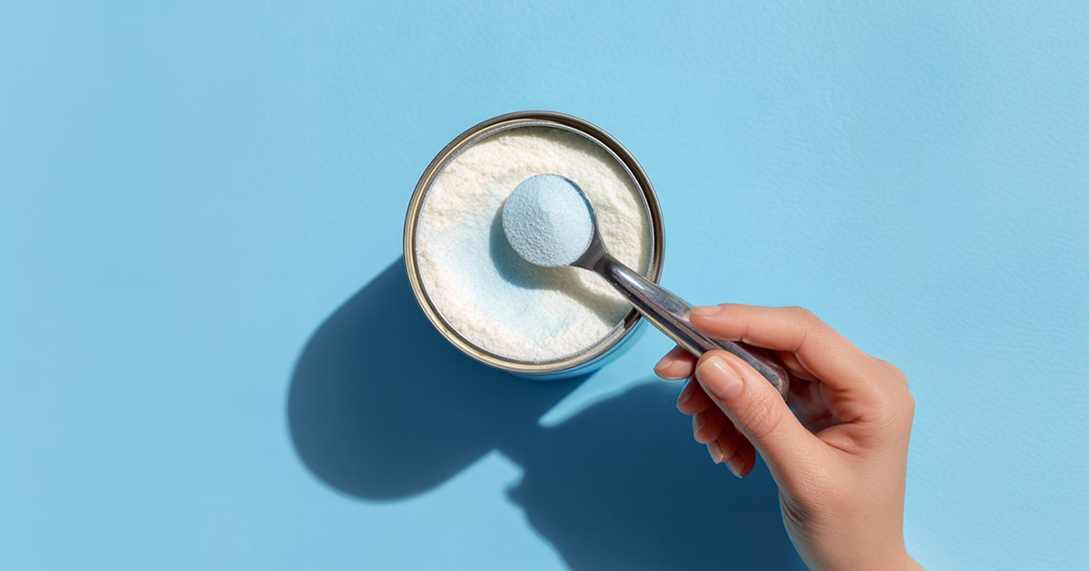 Hand holding a scoop of powdered baby formula over an open formula can on a blue background.
